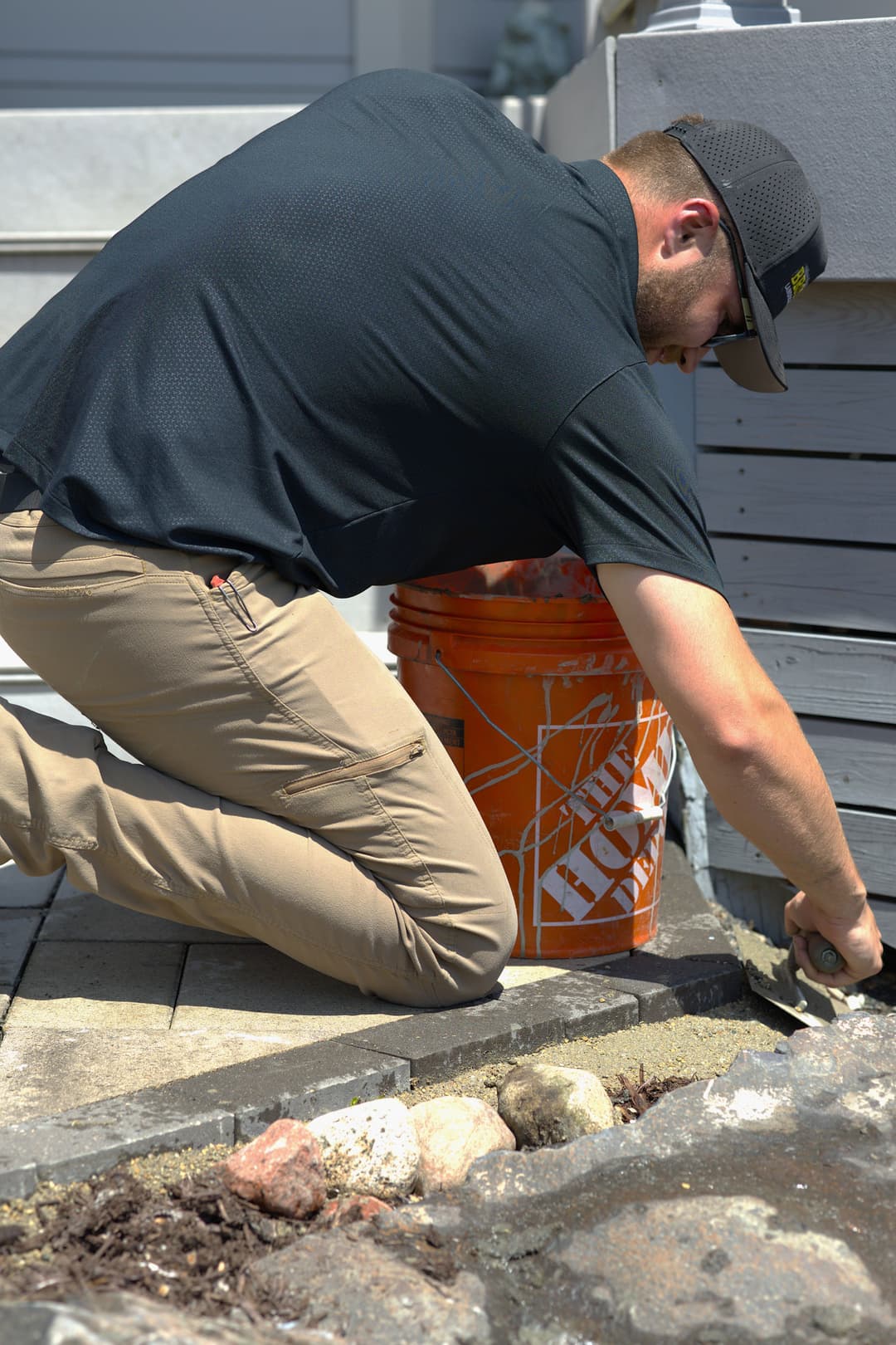 Man kneeling while laying concrete edge restraint along a paver walkway, with tools and a bucket visible.