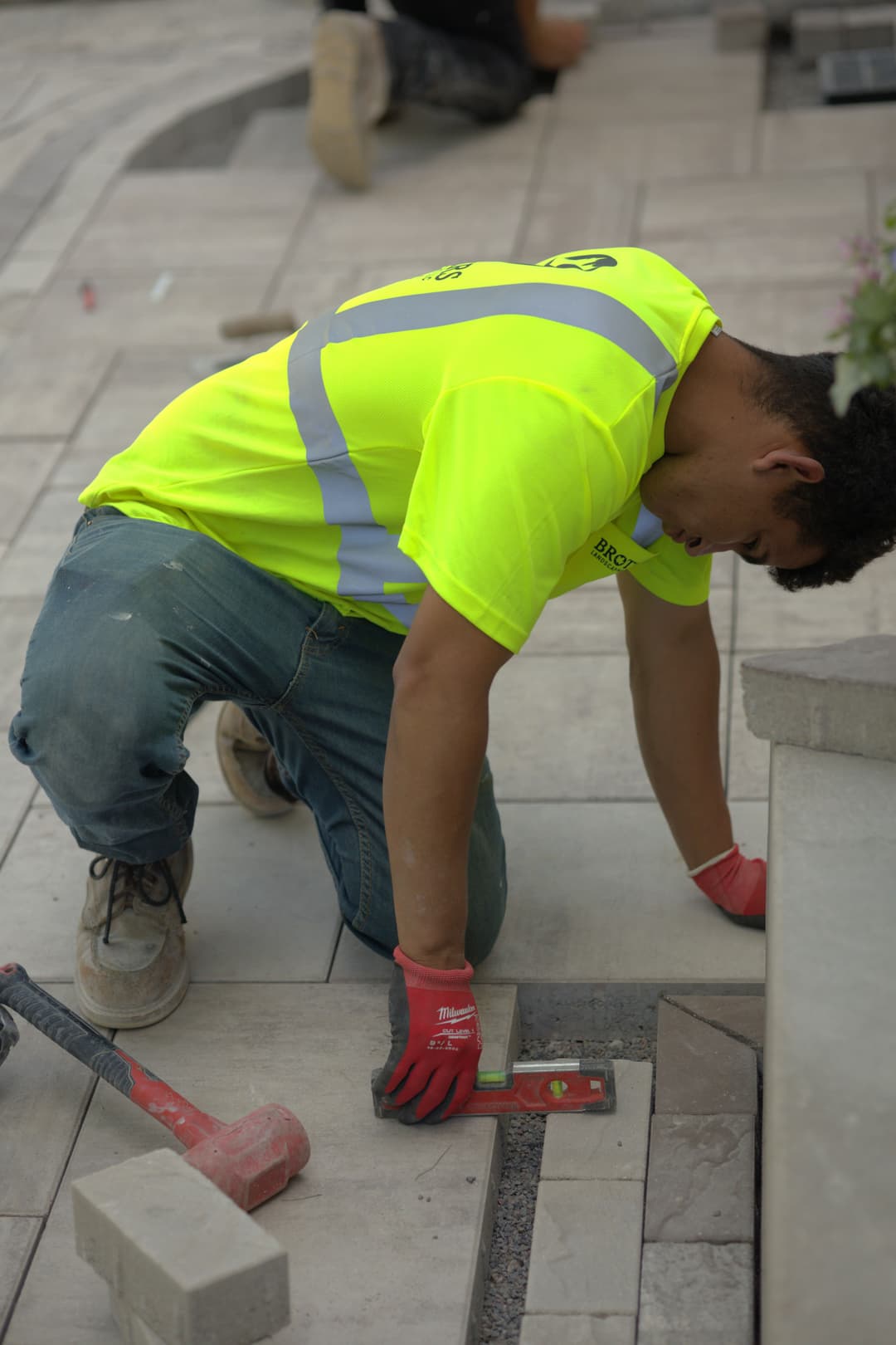 Worker laying pavers on a patio, wearing a neon yellow shirt and gloves, using a level tool.