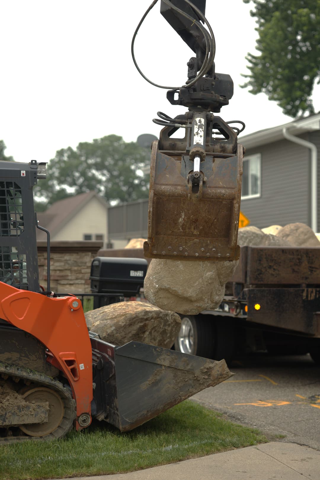 Excavator lifting large boulder at a construction site with a dump truck nearby.