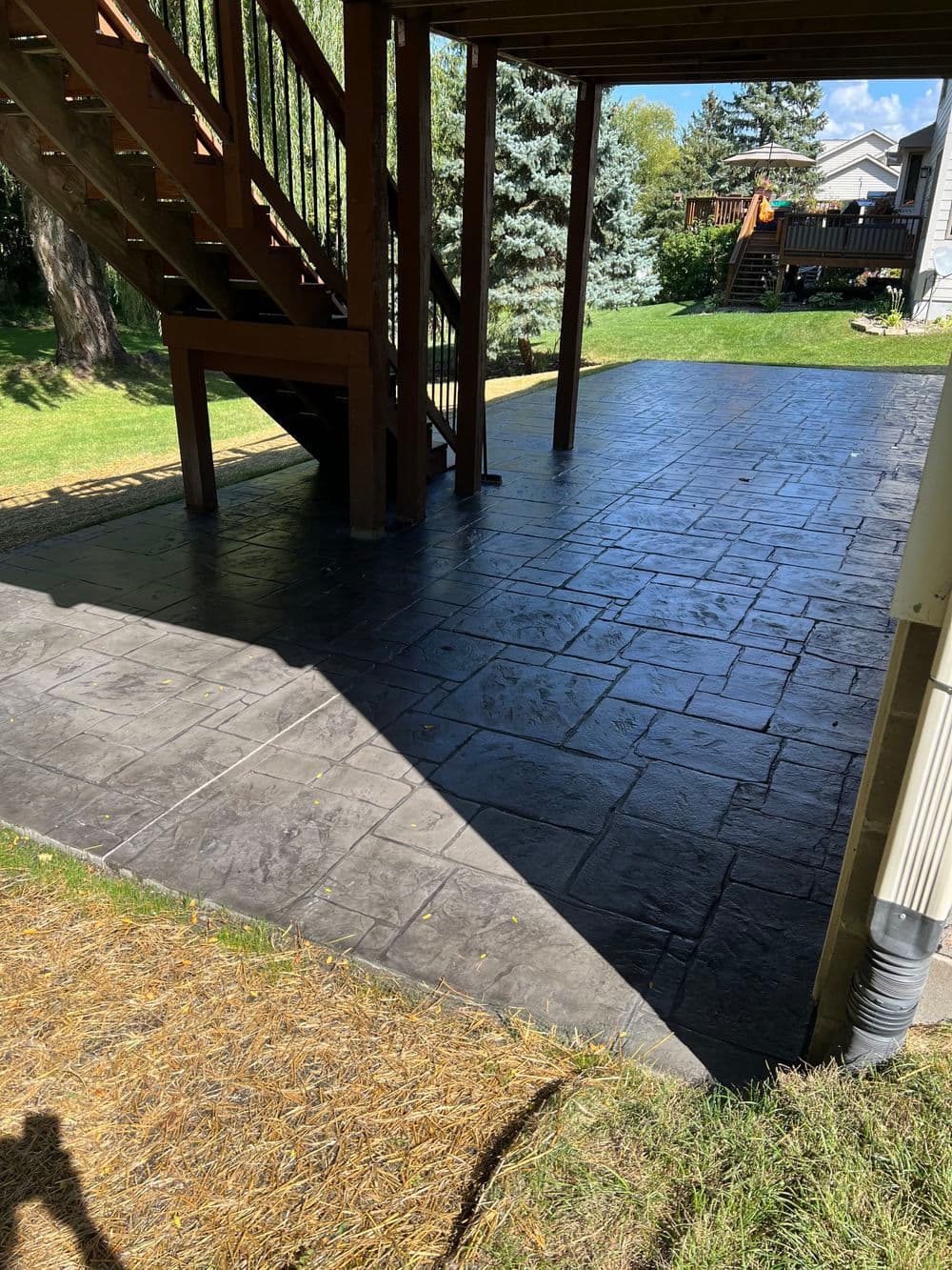Decorative stamped concrete patio under a wooden staircase with lush green surroundings.