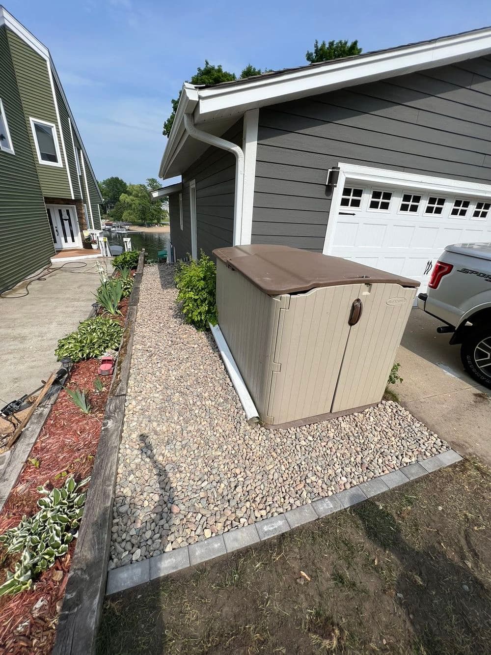 Storage shed on gravel pathway beside grey house with landscaped garden features.