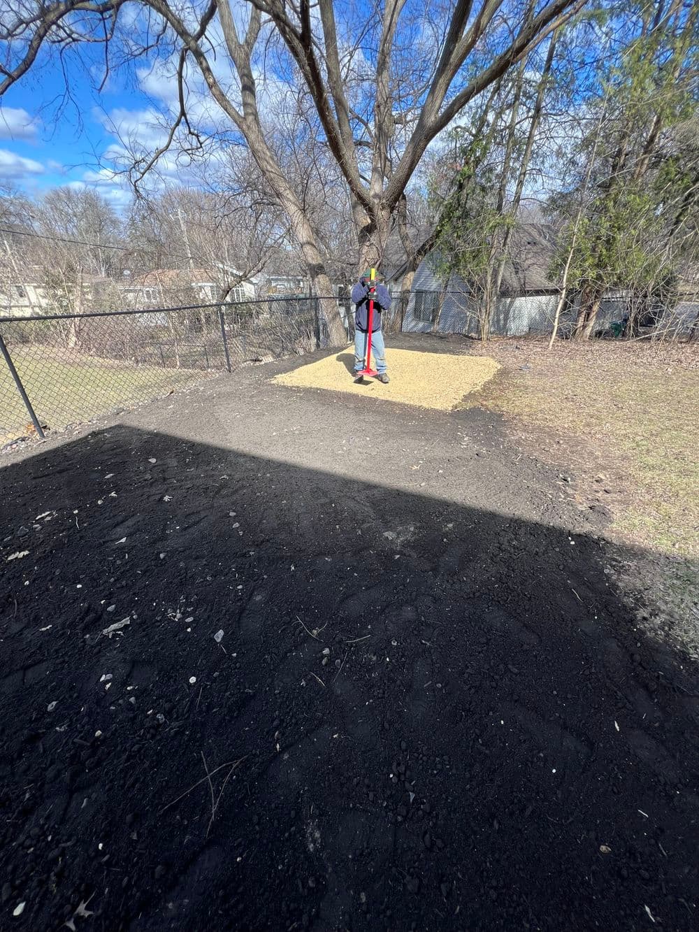 Person using a post-hole digger in a yard under a tree with a fenced area and clear sky.