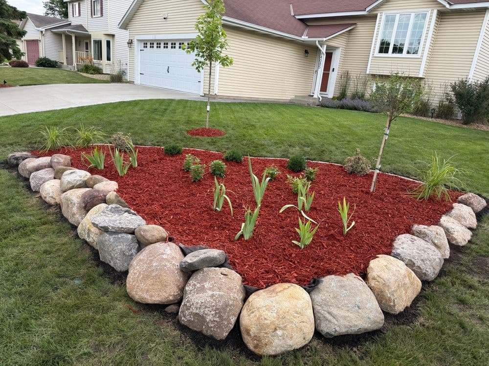 Red mulched flower bed with shrubs and plants surrounded by decorative stones in residential yard.