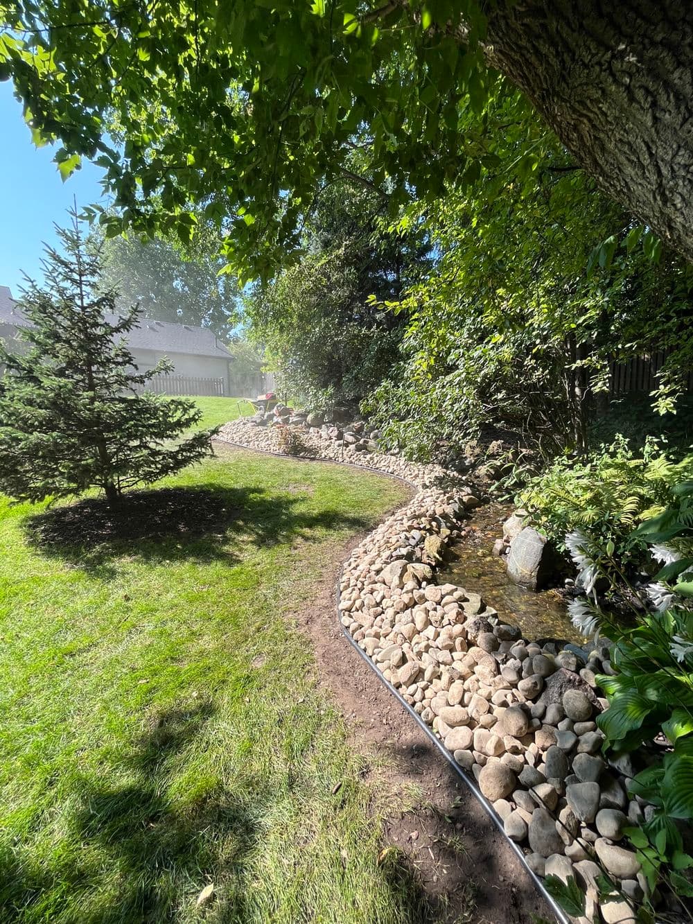 Lush garden pathway with rocks and stream surrounded by greenery and trees.