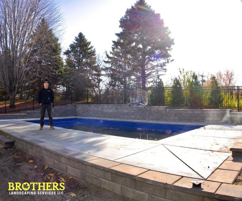 Landscaping professional stands by a newly constructed pool with stone patio and trees.