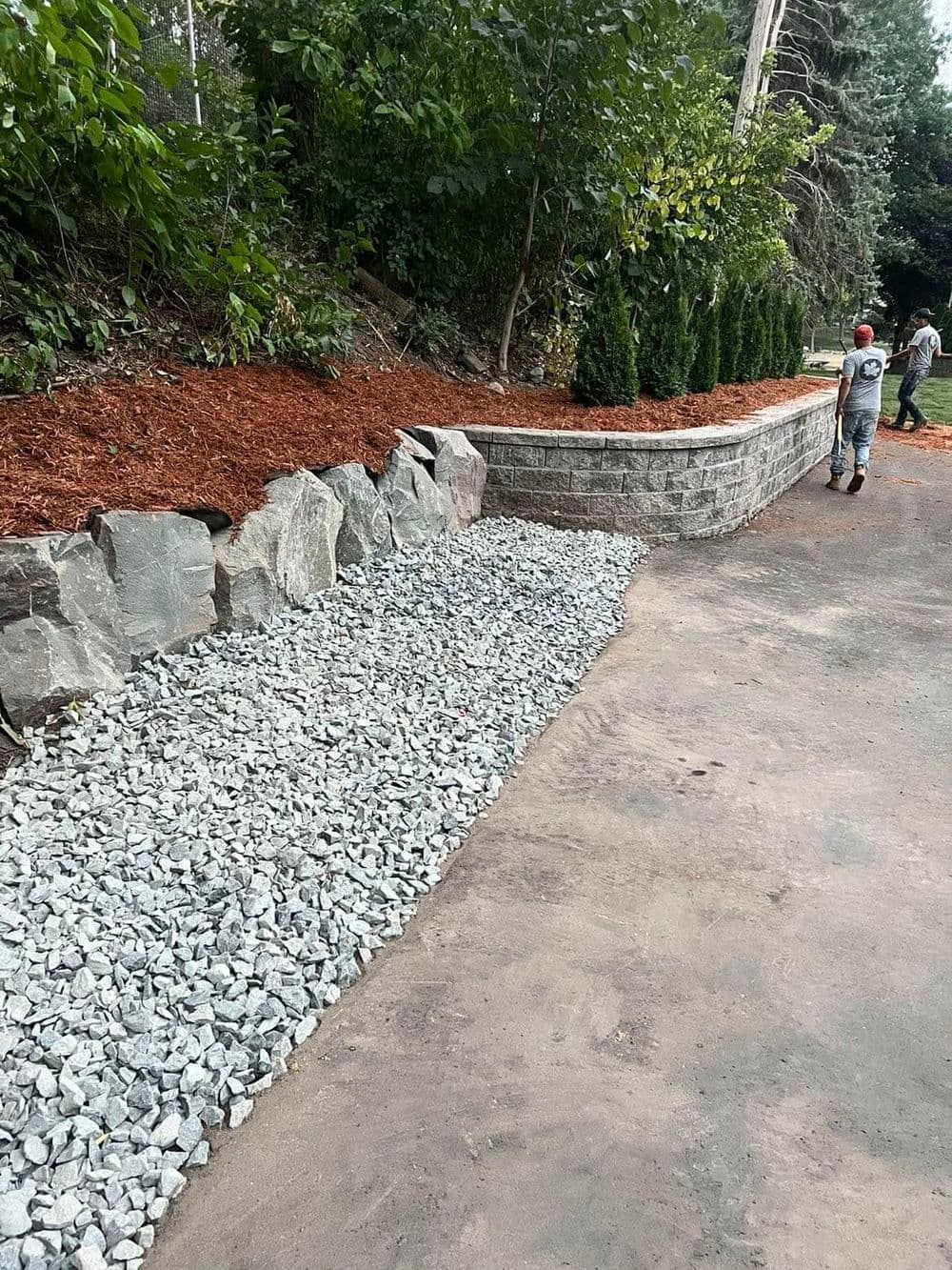 Landscaped path featuring gravel, stone wall, and mulch with greenery in the background.