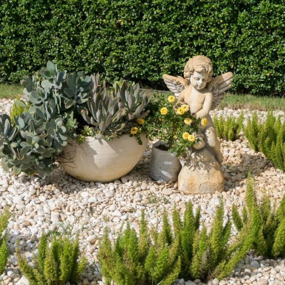 Stone angel statue surrounded by succulents and yellow flowers in a pebble garden.