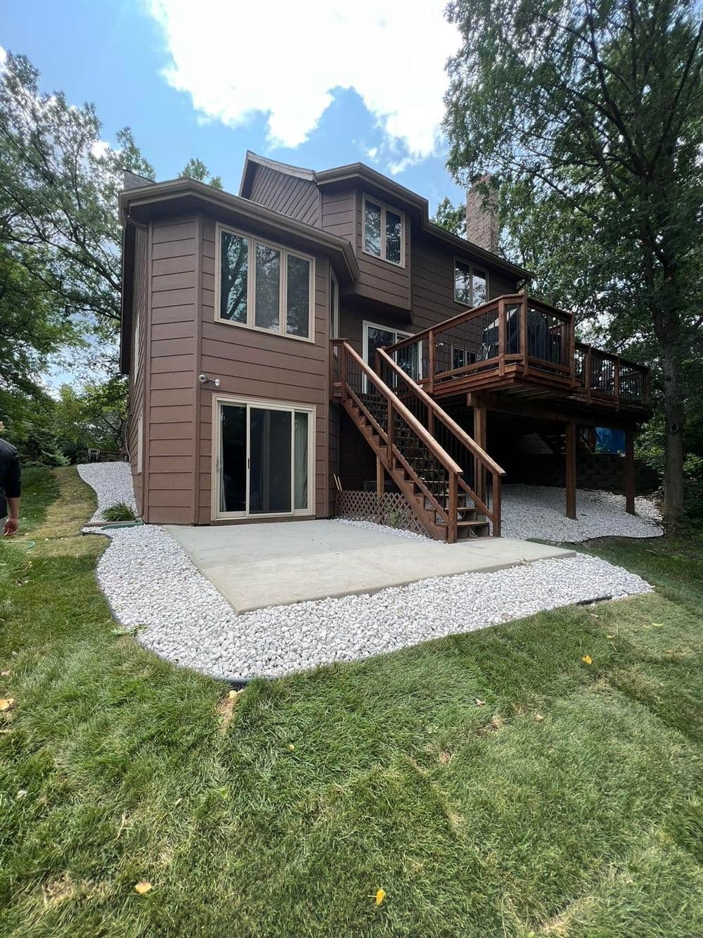 Two-story brown house with a wooden deck and stone landscaping in a green yard.