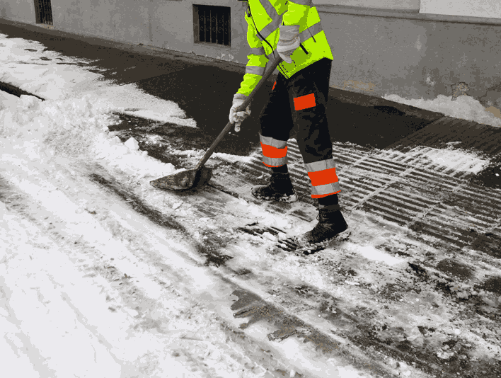 A man wearing a yellow jacket is actively shoveling snow off the sidewalk for clearing purposes.